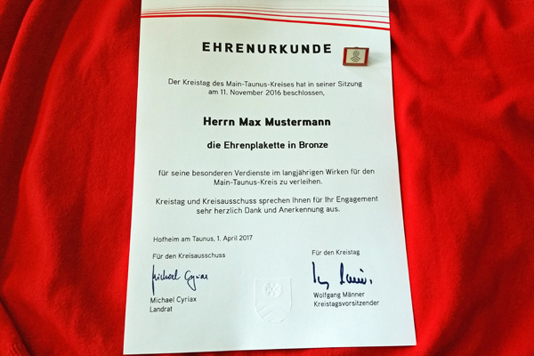 eine Urkunde über die Ehrenplakette in Bronze vor einem roten Hintergrund